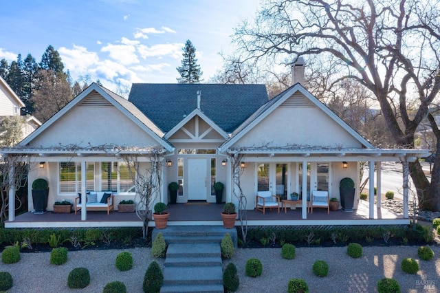 back of house with a porch, a shingled roof, a chimney, and stucco siding