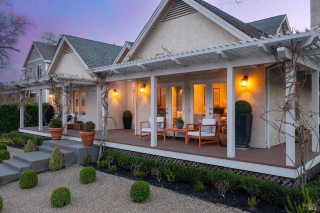 back of house at dusk featuring stucco siding, roof with shingles, a pergola, and french doors