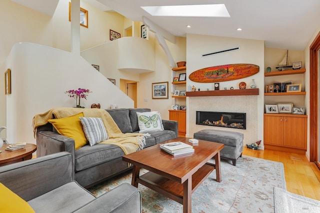 living room featuring high vaulted ceiling, a skylight, light wood-type flooring, built in shelves, and a fireplace