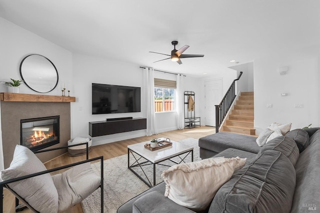 living area featuring stairs, a glass covered fireplace, a ceiling fan, and wood finished floors