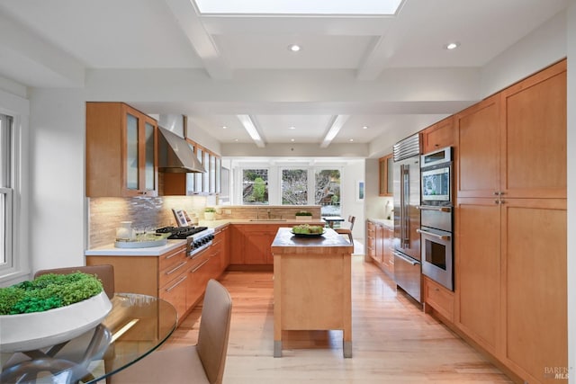 kitchen with a warming drawer, appliances with stainless steel finishes, glass insert cabinets, beamed ceiling, and wall chimney exhaust hood