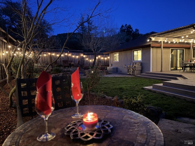 patio at night featuring a deck, outdoor dining space, a fenced backyard, and a fire pit