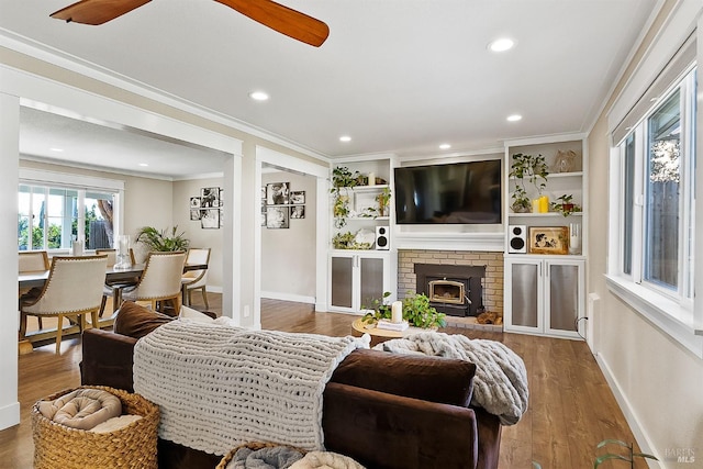 living room with baseboards, ceiling fan, wood finished floors, crown molding, and a brick fireplace