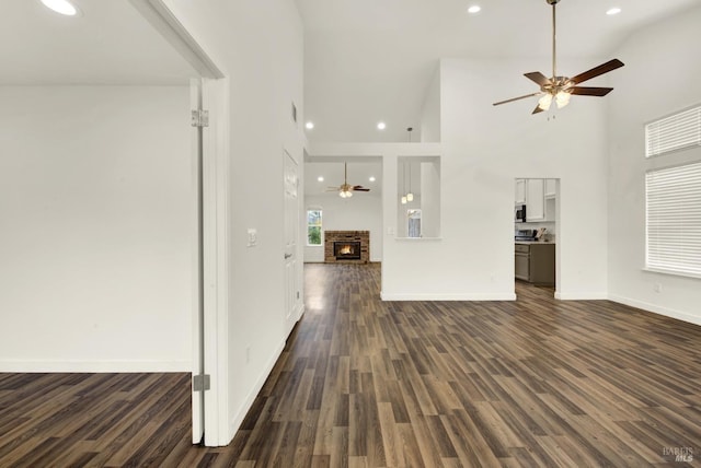 unfurnished living room with dark wood-type flooring, a ceiling fan, a warm lit fireplace, baseboards, and a towering ceiling