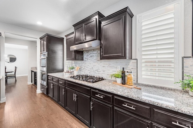 kitchen featuring stainless steel appliances, backsplash, light wood-style flooring, light stone countertops, and under cabinet range hood