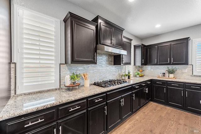 kitchen with stainless steel gas cooktop, light wood finished floors, decorative backsplash, light stone countertops, and under cabinet range hood