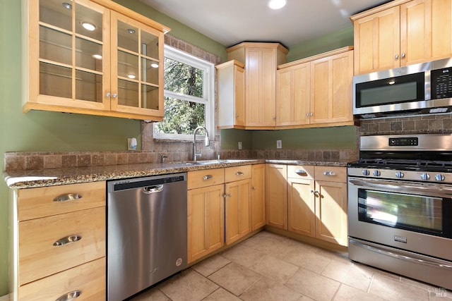 kitchen with light brown cabinets, stainless steel appliances, a sink, dark stone countertops, and glass insert cabinets