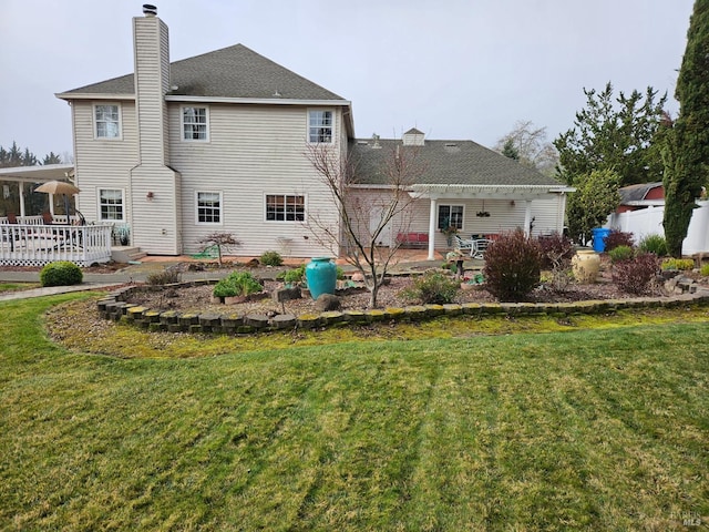 rear view of property featuring a patio area, a chimney, a deck, a shingled roof, and a lawn
