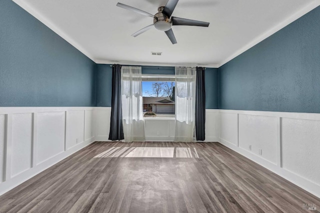 empty room with ceiling fan, visible vents, wood finished floors, and wainscoting