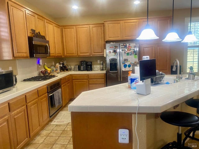 kitchen with a breakfast bar, an island with sink, tile countertops, stainless steel appliances, and hanging light fixtures