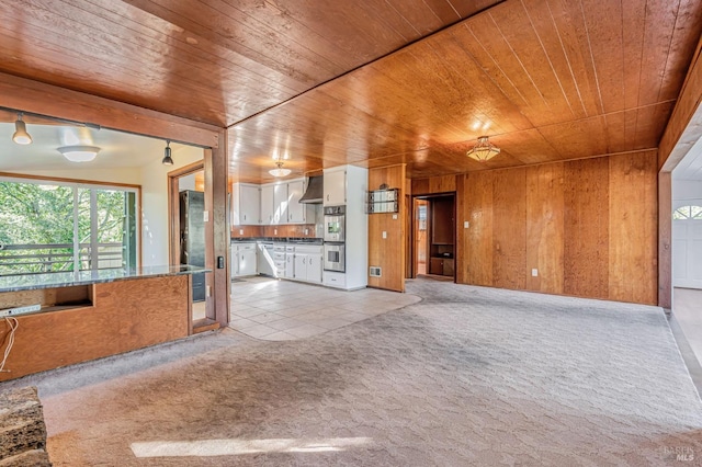 kitchen featuring wall chimney exhaust hood, light colored carpet, white cabinets, wooden ceiling, and wooden walls