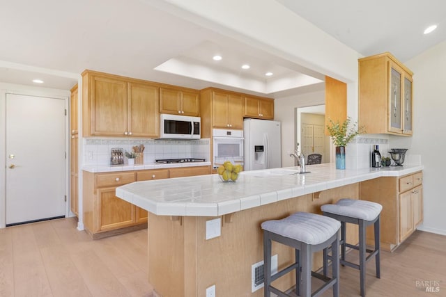 kitchen with a tray ceiling, light wood-style flooring, white appliances, a peninsula, and a kitchen bar