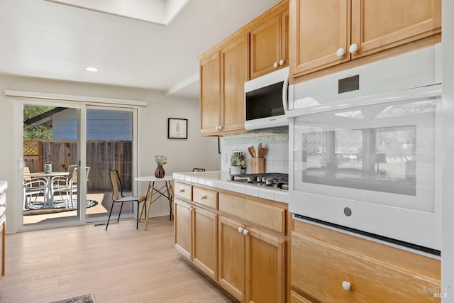 kitchen with light brown cabinets, light wood-style flooring, white appliances, backsplash, and tile counters