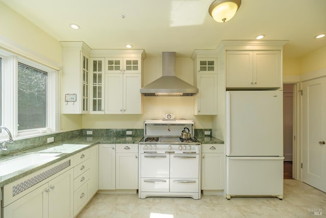 kitchen featuring wall chimney range hood, dark stone counters, white appliances, and a sink