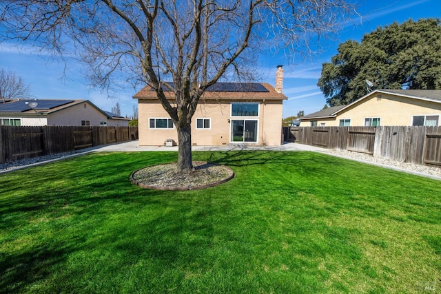 rear view of property with a patio, roof mounted solar panels, a fenced backyard, a yard, and a chimney