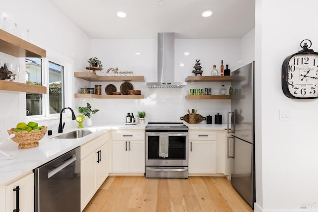 kitchen with wall chimney range hood, open shelves, appliances with stainless steel finishes, a sink, and light wood finished floors