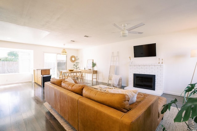 living room with visible vents, a ceiling fan, baseboards, a brick fireplace, and dark wood-style flooring