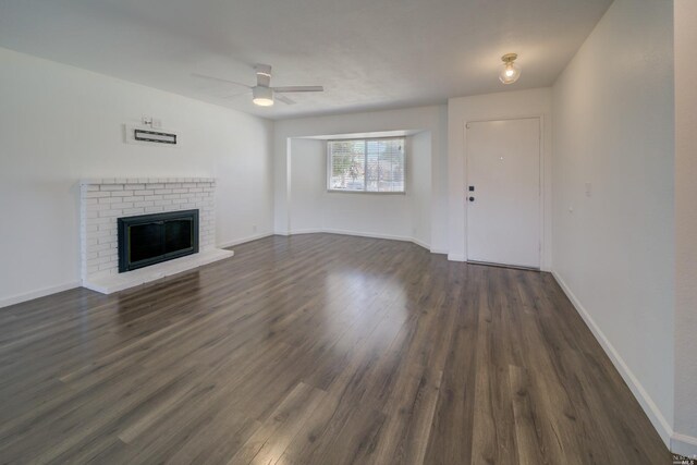 unfurnished living room with a brick fireplace, a ceiling fan, dark wood-style flooring, and baseboards