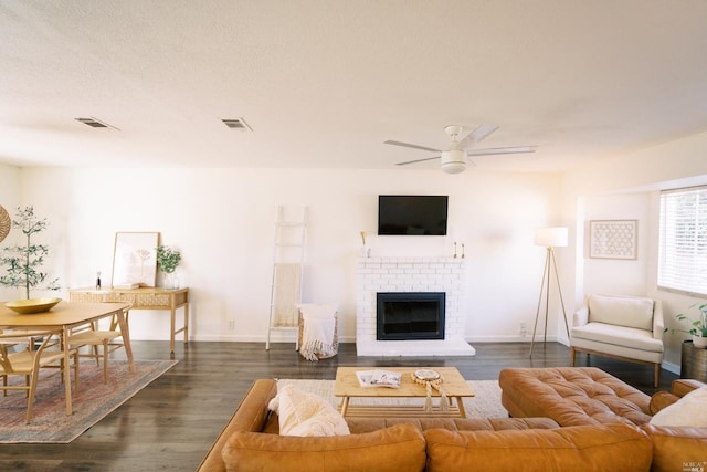 living room featuring visible vents, dark wood-type flooring, baseboards, a brick fireplace, and ceiling fan