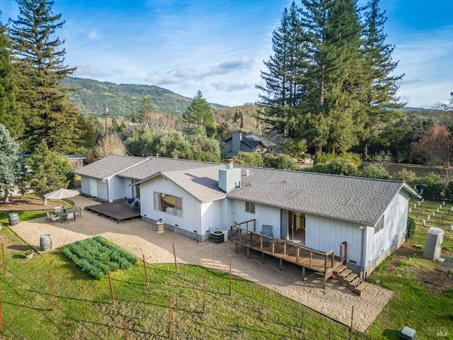 back of property with cooling unit, roof with shingles, a deck with mountain view, a chimney, and a patio area