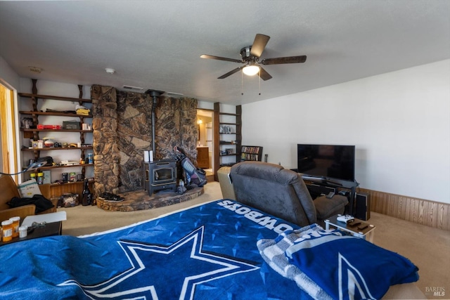 carpeted living room featuring wooden walls, built in shelves, a ceiling fan, a wood stove, and wainscoting
