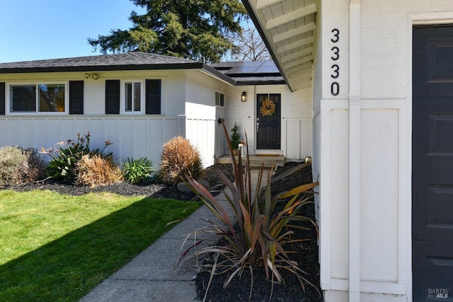 doorway to property featuring a lawn, roof mounted solar panels, roof with shingles, board and batten siding, and a garage