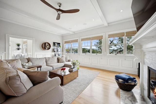 living area featuring beamed ceiling, a premium fireplace, wood finished floors, a decorative wall, and a ceiling fan