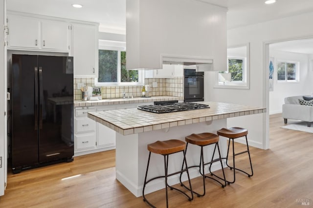 kitchen featuring tile countertops, light wood finished floors, decorative backsplash, black appliances, and a kitchen bar