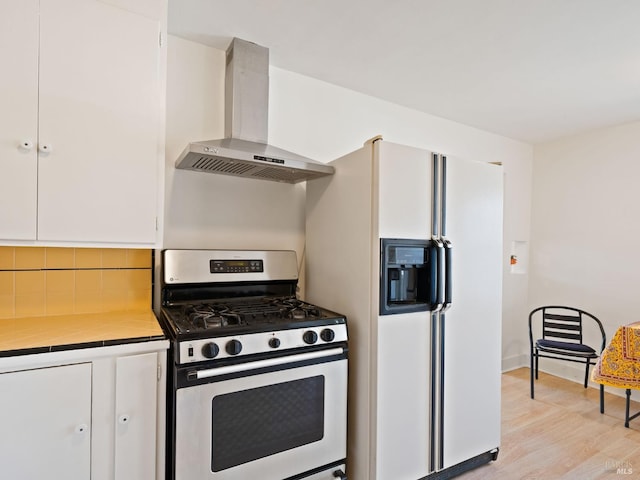 kitchen featuring tile countertops, white fridge with ice dispenser, white cabinetry, wall chimney range hood, and stainless steel gas stove