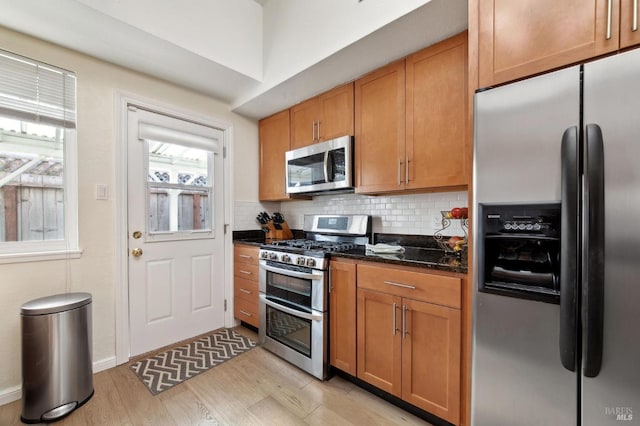 kitchen featuring dark stone countertops, brown cabinetry, light wood-style flooring, appliances with stainless steel finishes, and backsplash