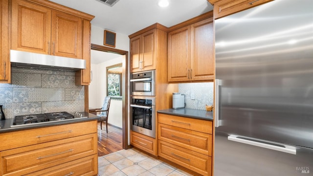 kitchen with under cabinet range hood, dark countertops, appliances with stainless steel finishes, brown cabinetry, and light tile patterned floors