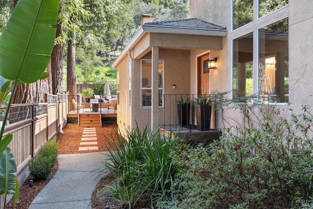 view of exterior entry with a shingled roof, fence, stucco siding, a chimney, and a deck