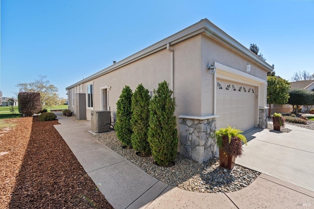 view of side of property featuring concrete driveway, stucco siding, central AC unit, a garage, and stone siding