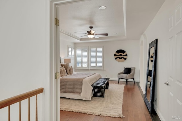 bedroom with ceiling fan, baseboards, a tray ceiling, wood finished floors, and arched walkways