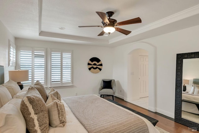 bedroom with baseboards, ceiling fan, wood finished floors, arched walkways, and a raised ceiling