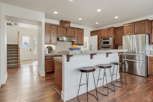 kitchen featuring dark wood finished floors, a breakfast bar, under cabinet range hood, appliances with stainless steel finishes, and backsplash