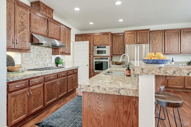 kitchen featuring under cabinet range hood, a sink, a kitchen breakfast bar, dark wood finished floors, and appliances with stainless steel finishes