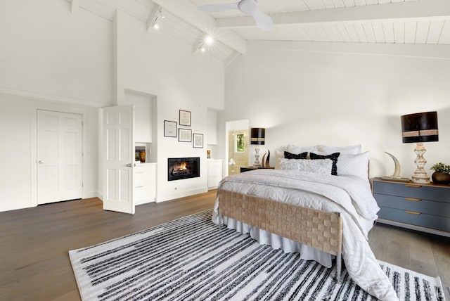 bedroom with beamed ceiling, high vaulted ceiling, track lighting, a glass covered fireplace, and wood finished floors