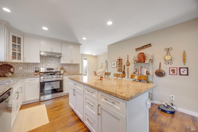 kitchen with under cabinet range hood, appliances with stainless steel finishes, white cabinetry, light wood-type flooring, and backsplash