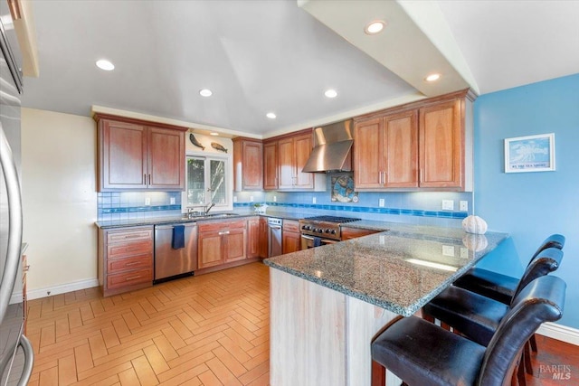 kitchen with dark stone countertops, a peninsula, stainless steel appliances, decorative backsplash, and wall chimney range hood