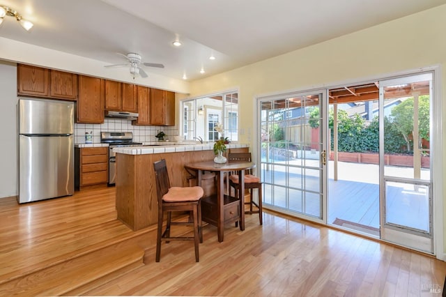 kitchen with backsplash, under cabinet range hood, light wood-style flooring, appliances with stainless steel finishes, and brown cabinetry