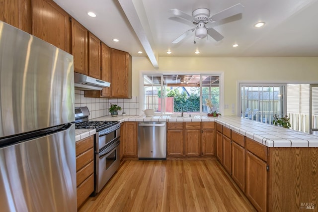 kitchen featuring under cabinet range hood, appliances with stainless steel finishes, a peninsula, light wood finished floors, and decorative backsplash