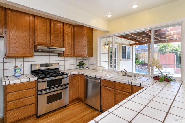 kitchen featuring under cabinet range hood, a sink, tile countertops, appliances with stainless steel finishes, and decorative backsplash