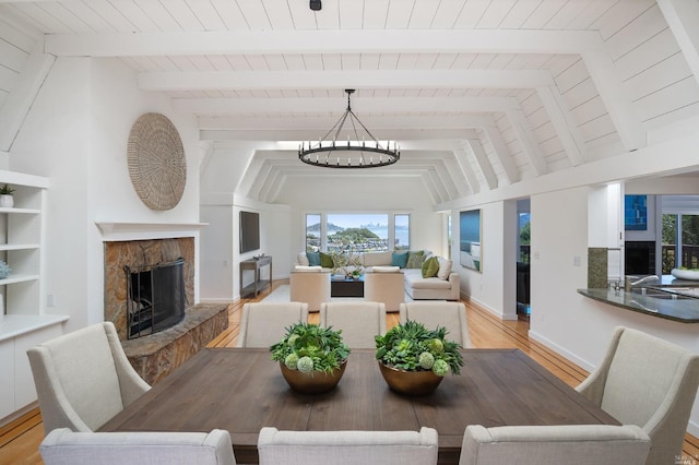 dining area with a notable chandelier, light wood-style flooring, a fireplace with raised hearth, lofted ceiling with beams, and baseboards