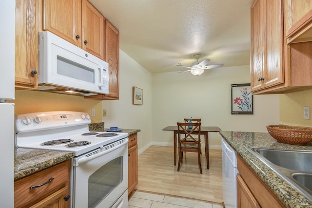 kitchen featuring baseboards, dark stone countertops, white appliances, a ceiling fan, and a sink