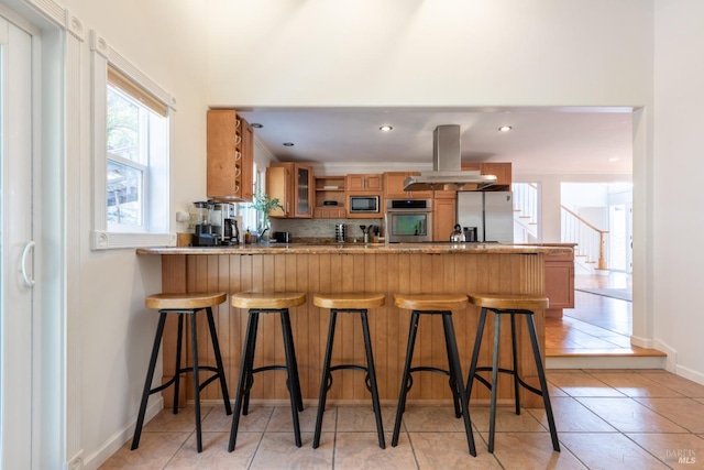 kitchen featuring brown cabinetry, a peninsula, light tile patterned flooring, stainless steel appliances, and island range hood