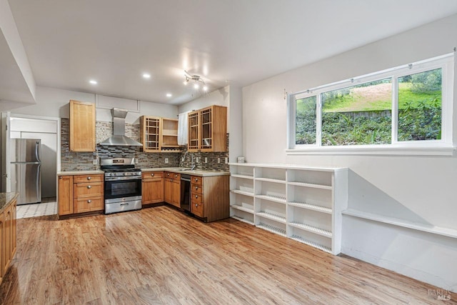 kitchen with light wood-type flooring, backsplash, stainless steel appliances, wall chimney range hood, and glass insert cabinets