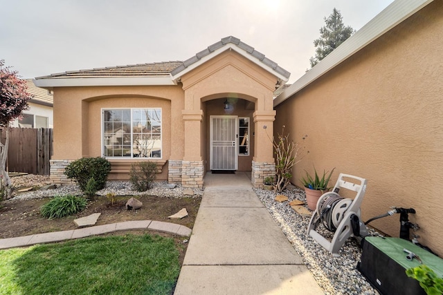 view of exterior entry with stucco siding, stone siding, a tile roof, and fence