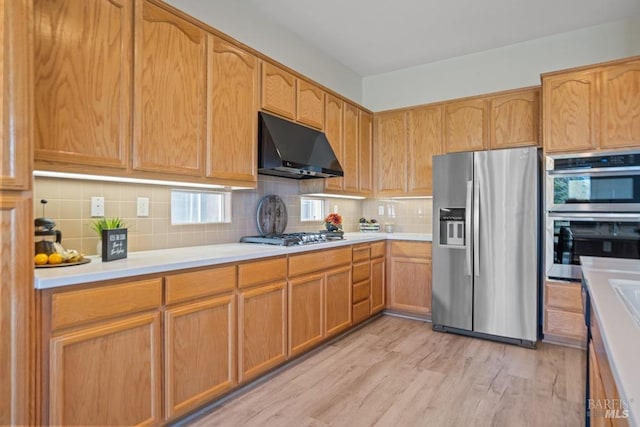kitchen with light wood-type flooring, light countertops, backsplash, under cabinet range hood, and appliances with stainless steel finishes
