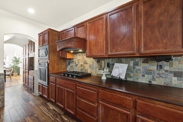 kitchen featuring dark countertops, backsplash, dark wood finished floors, arched walkways, and stainless steel appliances